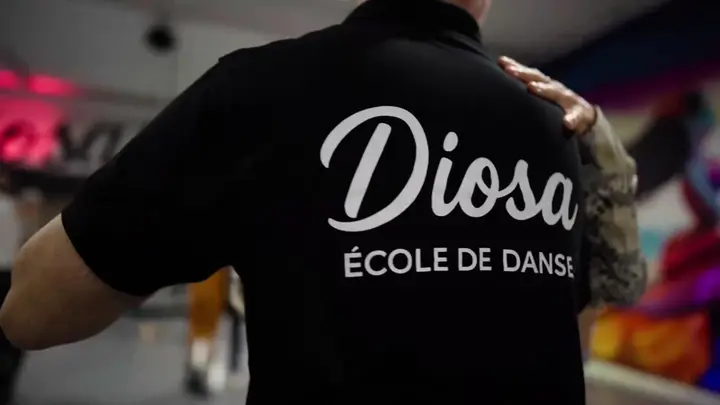 Danseur vu de dos en polo noir portant le logo « Diosa — École de danse », dans une salle de danse éclairée de néons