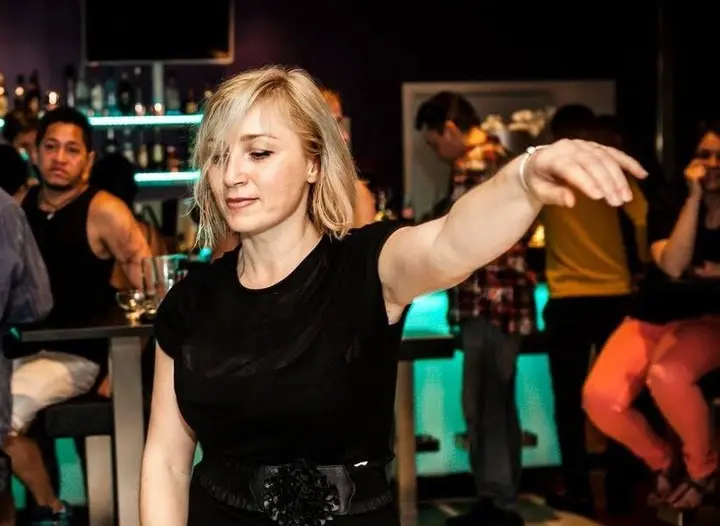 Photo d'ambiance d'une danseuse blonde vêtue de noir, bras tendu, sur une piste éclairée de turquoise devant le bar