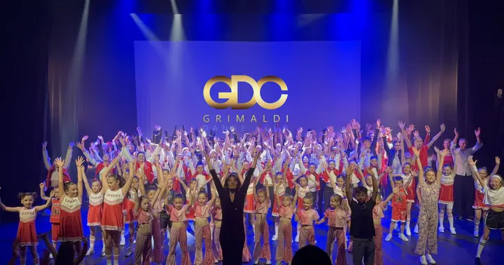 Photo de gala sur scène : élèves du Grimaldi Danse Club en costumes rouges et blancs, bras levés sous le logo doré GDC