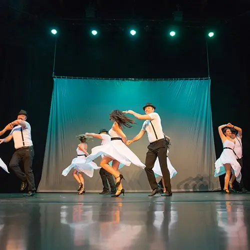 Photo de scène : des couples de danseurs en tenue blanche, hommes en chapeau et bretelles, femmes en robe tournoyante, devant un rideau turquoise.