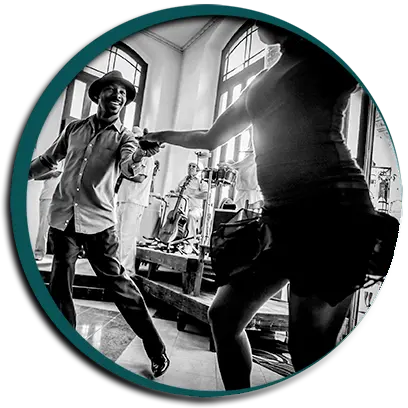 Photo en noir et blanc, dans un cercle bordé de vert, d'un danseur en chapeau qui sourit en guidant sa partenaire dans une salle où l'on voit des guitares.
