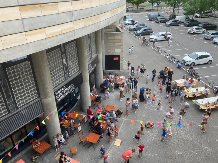 Vue plongeante sur le parvis de Malraux à Chambéry, tables de guinguette, guirlandes de fanions colorés et foule de familles.