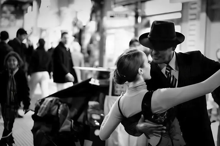 Photo en noir et blanc d'un couple enlacé dansant au milieu d'une rue passante, l'homme en costume et chapeau feutre.