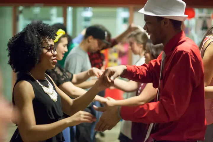 Photo d'une piste de danse : un homme en chemise rouge et chapeau blanc guide sa partenaire en robe noire, d'autres couples dansent derrière eux.