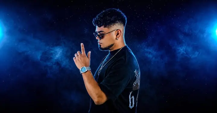 Photo d'un jeune homme de profil en t-shirt noir et lunettes de soleil, l'index levé, sur fond bleu étoilé, sans aucun texte.