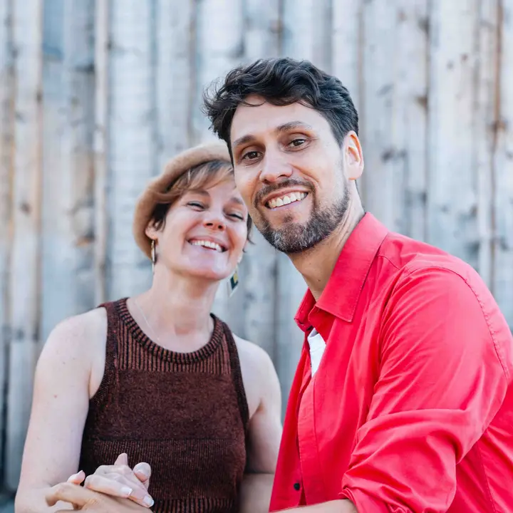 Portrait souriant d'Aude & Pom, joue contre joue devant une palissade de bois clair, elle en béret beige et lui en chemise rouge.