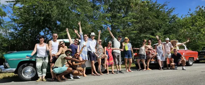 Photo de groupe de danseurs souriants, bras levés, devant des voitures américaines vintage turquoise et rouge sous le soleil cubain.